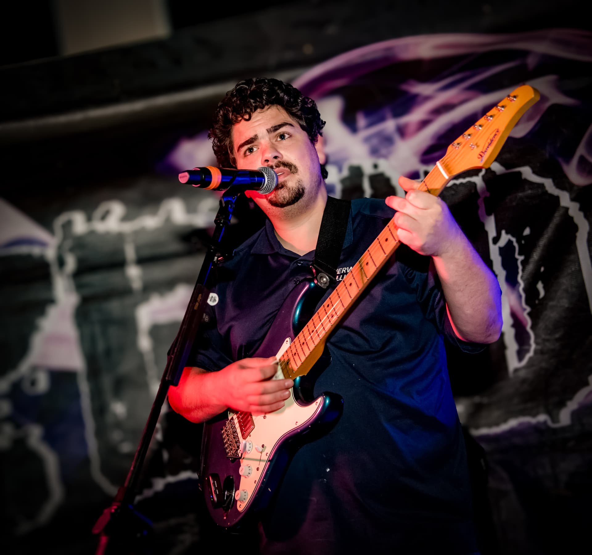 Jake Frezza singing into a microphone while playing electric guitar under stage lights